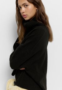 Črn pulover s turtleneck ovratnikom, ki ima rebrasto ovratnik in manšete, narejen iz mehkega pletenega materiala, z sproščenim krojem in minimalnimi detajli.