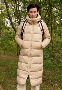 Cappotto lungo imbottito beige con zip frontale, colletto alto, tasche laterali e fondo spaccato, indossato con spalline nere, ambientato in una foresta verde.