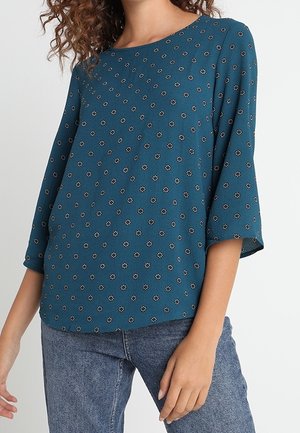 Frau trägt ein türkisblaues Langarmshirt mit kleinen weißen Blumenmustern und hellblaue Jeans, steht mit einem leicht ausgestreckten Arm.