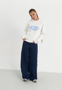 Felpa bianca con testo blu "Fai ciò che ti rende felice", abbinata a jeans a gamba larga in denim scuro e sneakers. Design semplice e casual.