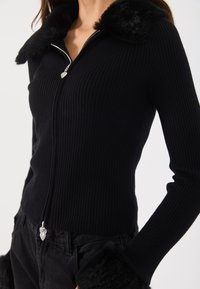 Czarny prążkowany sweter z długim rękawem z sztucznym futerkiem przy kołnierzu i mankietach, z zamkiem błyskawicznym w kolorze srebrnym i suwadłem w kształcie serca.