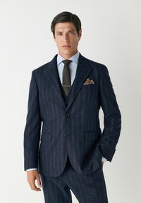 Sötétkék csíkos öltöny, két gombos zárással, hozzá illő nadrággal és világoskék inggel. Mintás nyakkendő és zsebkendő is van hozzá.