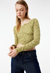 Grünes, kurz geschnittenes Oberteil mit strukturiertem Muster und langen Ärmeln, das über einen quadratischen Ausschnitt und eine figurbetonte Passform verfügt. Kombiniert mit dunklen Jeans.