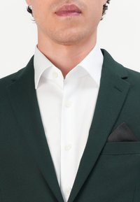 Dark green blazer with a white shirt underneath, featuring a black patterned pocket square and visible button details on the shirt.