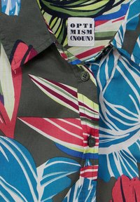 Camisa de manga corta con botones en tela gris oscuro con un vibrante patrón floral. Presenta una etiqueta blanca con la inscripción "OPTIMISMO" y cierre de botones negros.