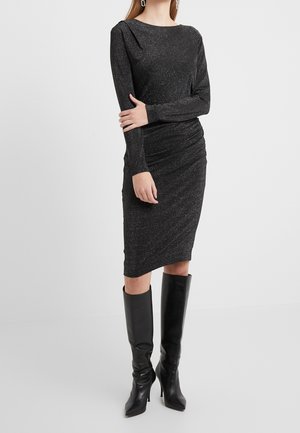 Mujer con un vestido negro ajustado de manga larga y largo hasta la rodilla con brillo metálico y botas negras de tacón hasta la rodilla, de pie frente a un fondo sencillo.