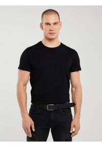 Homme aux cheveux courts portant un t-shirt noir et un jean noir avec une ceinture noire dans un studio au décor neutre.