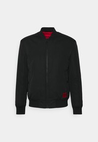 Blouson aviateur noir avec doublure rouge, fermeture éclair, poignets et ourlet côtelés, poches latérales et une petite étiquette logo rouge en bas à gauche.