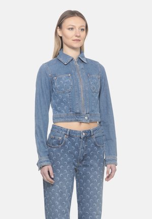 Donna che indossa una giacca di jeans corta abbinata e jeans a vita alta con motivi di luna crescente su sfondo grigio chiaro.