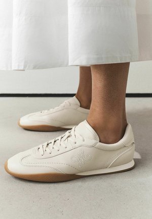 Person iført cremefarvede snøresneakers med gummisål og hvide viddebukser står på et lyst gulv.