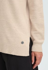 Beige džemperis, izgatavots no mīksta adījuma materiāla, ar ribotām piedurknēm un sānu šķirbām. Apakšējā kreisajā stūrī ir mazs, apaļš emblēma.