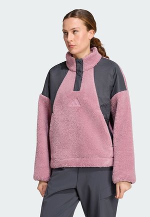 Mujer con un forro polar rosa y gris con cuello alto y cremallera parcial, combinado con pantalones gris oscuro, de pie frente a un fondo liso.