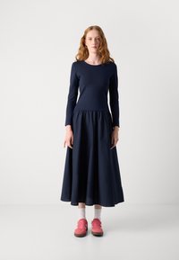 Mekko, jossa on pitkät hihat, tummansininen väri, kerätty vyötärö, laaja helma ja yhtenäinen rakenne. Yhdistetty vaaleanpunaisiin lenkkareihin ja valkoisiin sukkiin.