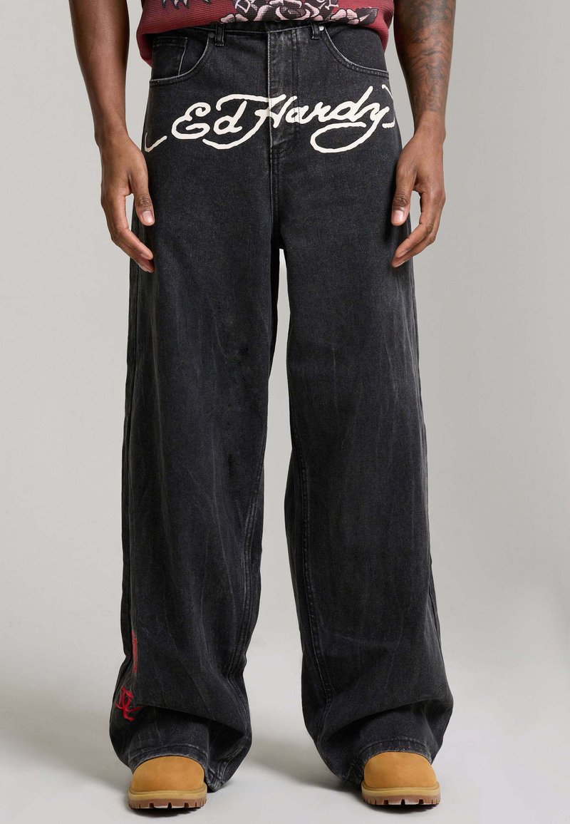 Jeans neri a gamba larga con la scritta bianca "Ed Hardy" sulla parte anteriore della vita, indossati con stivali color ocra e un braccio tatuato visibile.