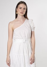 Vestido blanco de un solo hombro con manga abullonada, cintura ceñida y cinturón decorativo. Presenta detalles florales bordados en la falda acampanada.