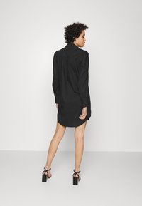 Vestido preto com colarinho, mangas longas e um corte solto. Apresenta botões ao longo das mangas e uma bainha ligeiramente curva.