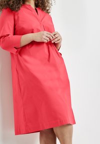 Žena se opírá o zeď a má na sobě volné korálové šaty ke kolenům s tříčtvrtečními rukávy a výstřihem do V.