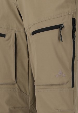 Tan cargobyxor med flera dragkedjefickor och svarta dragkedjehandtag, som visar detaljerad sömnad och tygtextur.