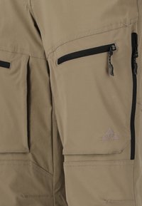 Pantalons d'extérieur beiges avec poches zippées noires, tissu renforcé et logo brodé sur la jambe inférieure. Design durable et léger.