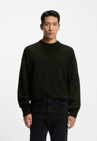 Dunkelgrüner Strickpullover mit Rundhalsausschnitt und langen Ärmeln, mit lockerer Passform und gerippten Bündchen, kombiniert mit schwarzen Jeans.