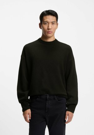 Dunkelgrüner Strickpullover mit Rundhalsausschnitt und langen Ärmeln, mit lockerer Passform und gerippten Bündchen, kombiniert mit schwarzen Jeans.