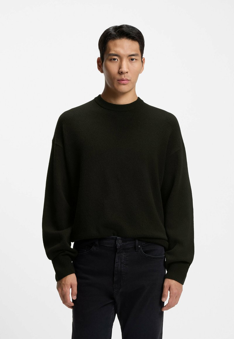 Dunkelgrüner Strickpullover mit Rundhalsausschnitt und langen Ärmeln, mit lockerer Passform und gerippten Bündchen, kombiniert mit schwarzen Jeans.