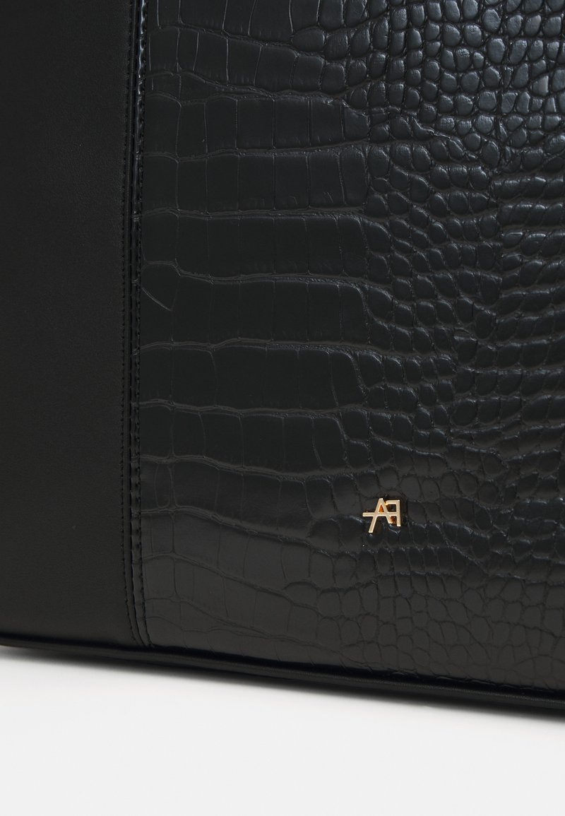 Sac à main noir en cuir lisse et texturé avec un motif crocodile. Accent logo doré sur le devant. Design élégant et minimaliste.