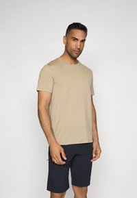 Camiseta de mangas cortas color beige en tela suave, con cuello redondo, combinada con pantalones cortos oscuros. El modelo adopta una postura neutral frente a un fondo claro.