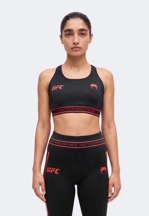 Femme portant un soutien-gorge de sport et un legging noirs UFC avec un texte rouge "Fight For Greatness", debout face à un fond uni.