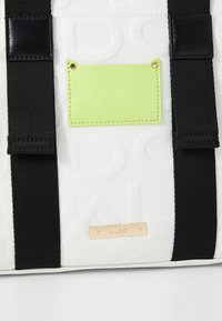 Weiße Tasche mit geprägtem Muster, schwarzen Riemen, goldfarbener Logo-Plakette und einem kleinen grünen Anhänger mit strukturierter Oberfläche.