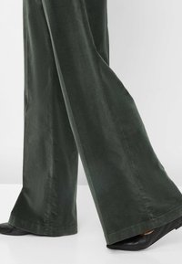 Pantalons larges vert foncé en tissu doux, avec une texture lisse et un ourlet net, associés à des chaussures noires à bout pointu.