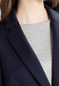 Námořnický modrý blazer se strukturovanými klopy, vyrobený z texturované látky. Nosený přes světle šedý top, který ukazuje přiléhavý střih u výstřihu.