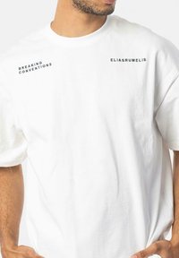 White cotton t-shirt with short sleeves. Features black text "BREAKING CONVENTIONS" and "ELIASRUMELIS" on the front. Simple, relaxed fit.