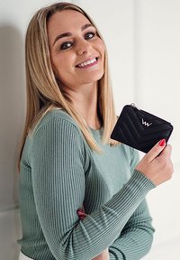 Black chevron-patterned wallet with silver hardware held in a hand, alongside a light green ribbed sweater, set against a neutral background.