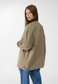 Overdimensioneret beige jakke med en mørkebrun krave, der har en knaplukning og sidelommer, parret med lyseblå jeans.