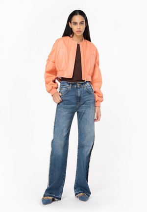 Giacca bomber arancione in pelle, tagliata, con zip, sopra marrone e jeans blu a gamba larga con fessure laterali e bordi sfrangiati.