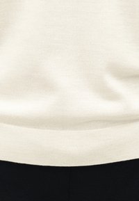 Πλεκτό πουλόβερ σε κρεμ χρώμα με ριμπ τελείωμα, φορεμένο πάνω από μαύρο παντελόνι, που αποκαλύπτει λεπτομέρειες στην περιοχή της μέσης και την υφή του υφάσματος.