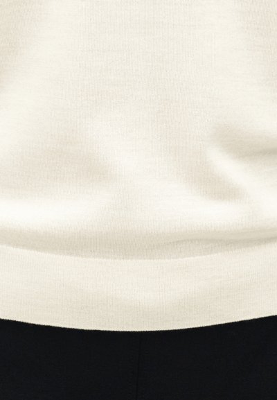 Pull en tricot couleur crème avec ourlet côtelé porté sur un pantalon noir, montrant la section médiane et les détails de la texture du tissu.