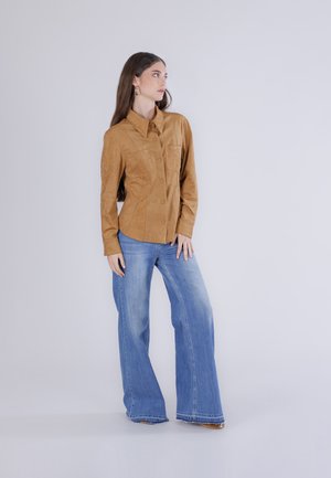 Mujer de pie, con una camisa marrón abotonada y pantalones vaqueros azules de pierna ancha, mirando hacia su izquierda contra un fondo claro y liso.