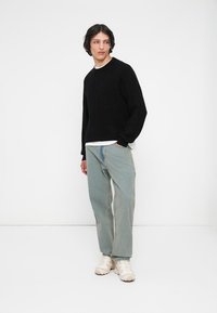 Ung mann står mot en hvit vegg iført sort genser, lyseblå jeans og hvite sneakers, og ser til venstre.