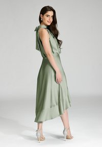 Robe verte au toucher soyeux avec une silhouette fluide, un ourlet volé et un détail en nœud à l'arrière. Associée à des talons argentés à lanières.