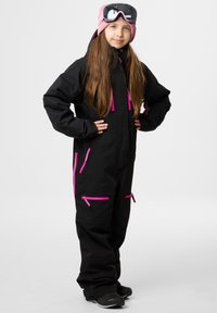 Zwarte sneeuwoutfit met roze ritsen en accenten, voorzien van een capuchon, meerdere ritszakken en een ontspannen pasvorm. Gediende met een skibril en een roze beanie.