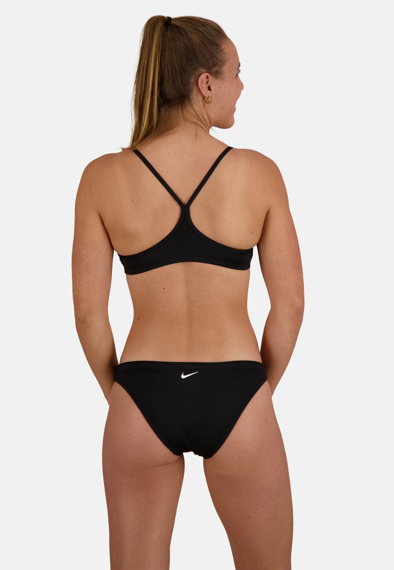 Czarny bikini z designem typu racerback i cienkimi ramiączkami. Posiada białe logo Nike na biodrze, gładką fakturę i minimalne pokrycie.