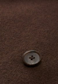 Bruine plastic knop met vier gaten, geplaatst op een gestructureerde donkerbruine stofachtergrond. Cirkelvormig met een gladde afwerking.