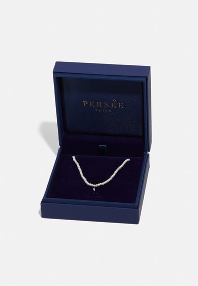 Pērļu kaklarota, kas ievietota zīdā izklātā tumši zilā kastē, ar maziem apaļiem pērļiem un vienkāršu piekariņu elegantiem akcentiem. Iekšpusē ir iegravēts zelta logo.