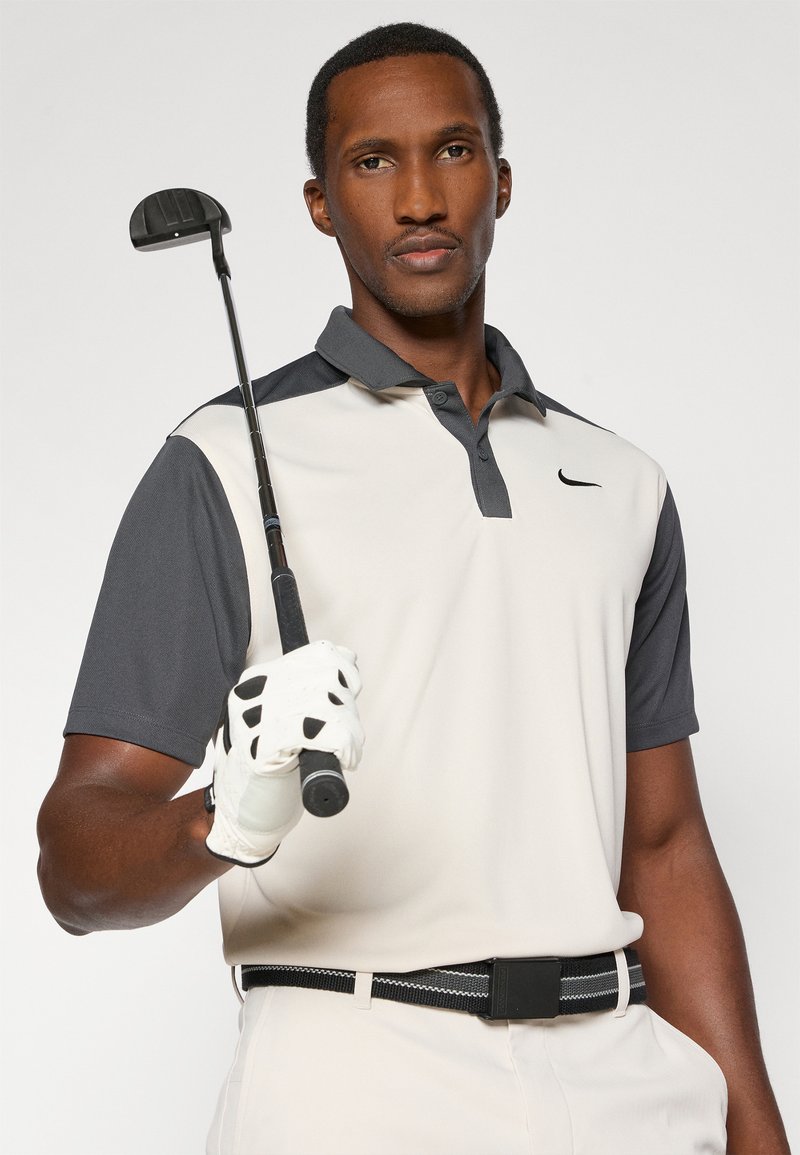 Hemd mit kurzen Ärmeln in einem hellbeige Farbton und dunklen grauen Ärmeln, mit einem Kragen und subtilem Nike-Logo. Hält einen Golfschläger und trägt einen weißen Handschuh.