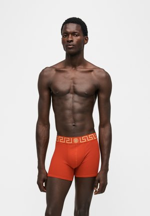 Modèle masculin à la peau foncée portant un boxer orange avec une ceinture à motif clé grecque et un médaillon central sur un fond blanc uni.