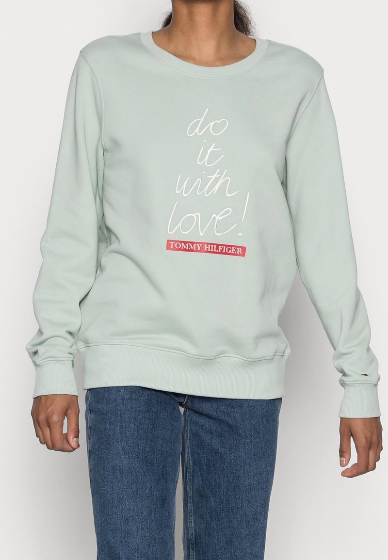 Sudadera verde claro con el texto "¡hazlo con amor!" y el logo de Tommy Hilfiger, llevada con jeans azules, mostrada de cuello a caderas.