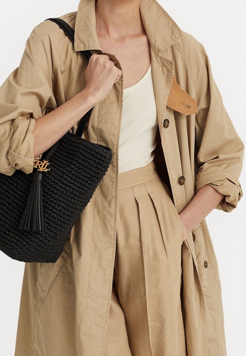 Personne portant un trench beige et un pantalon plissé, tenant un sac à main tissé noir avec un détail de pompon sur l'épaule.
