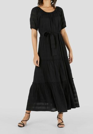 Femme portant une robe longue noire à étages, manches courtes, col rond et ceinture à la taille, associée à des sandales noires à brides.